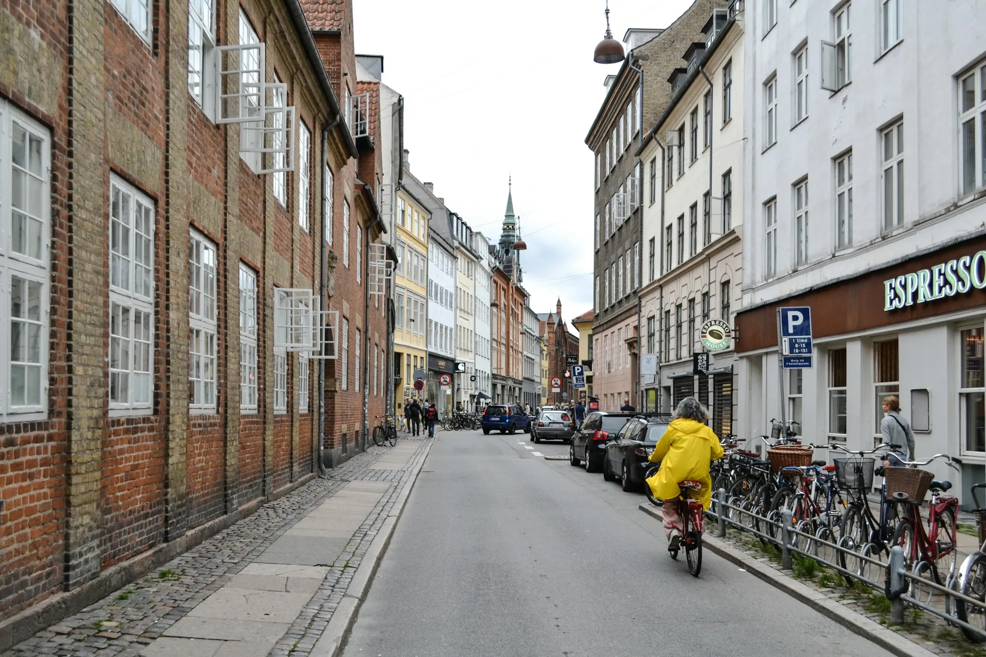 A cyclist in a yellow raincoat riding down a cobbled Copenhagen street lined with bicycles and cafés