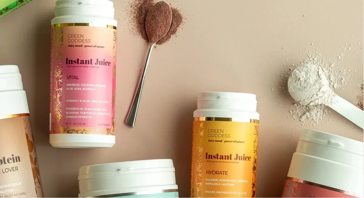Green Goddess Instant Juice and protein tubs on a pink table, with spoons of juice and protein powder