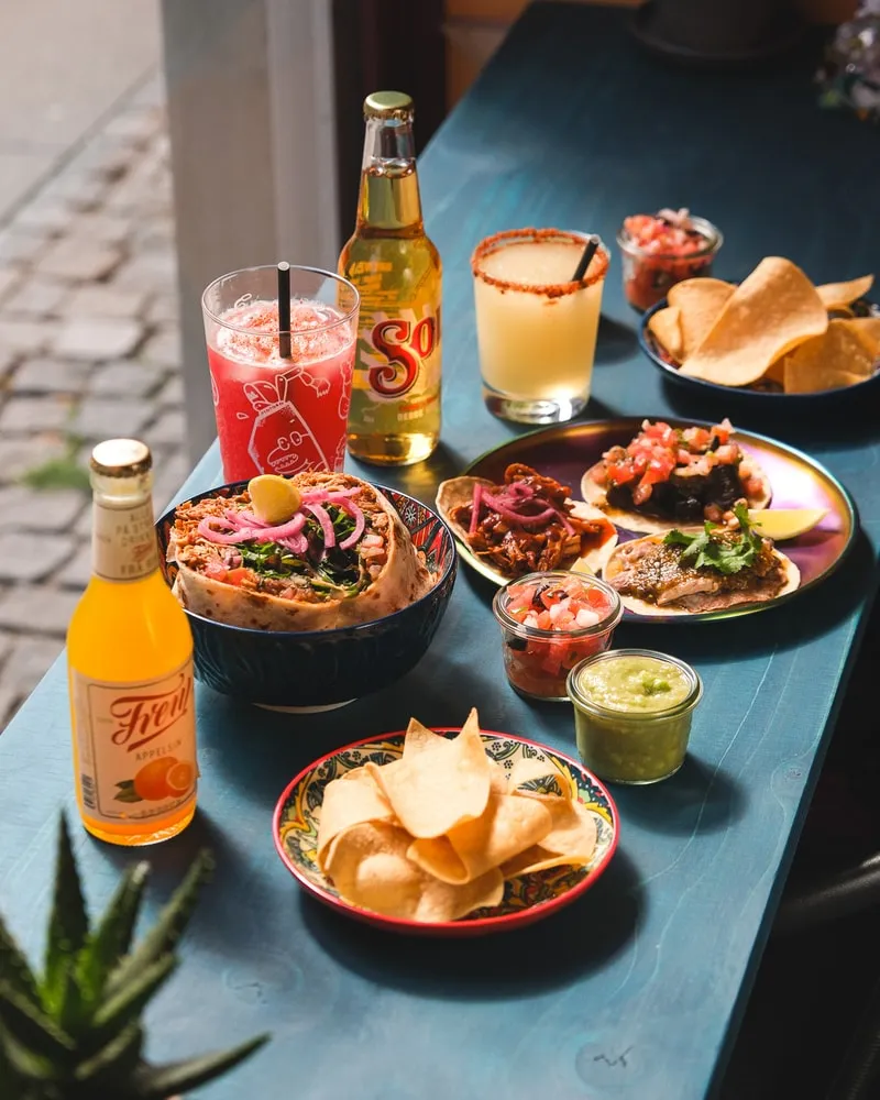 Tacos, a burrito bowl, chips and salsas with a margarita and cold drinks on a blue table by the window at Poco Loco