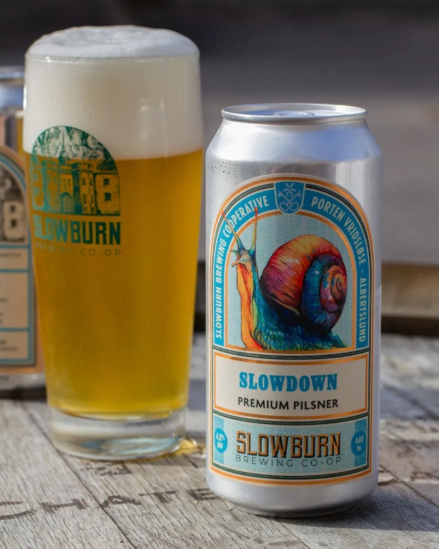 A can of Slowburn Slowdown premium pilsner beside a full glass of beer on a weathered wooden table