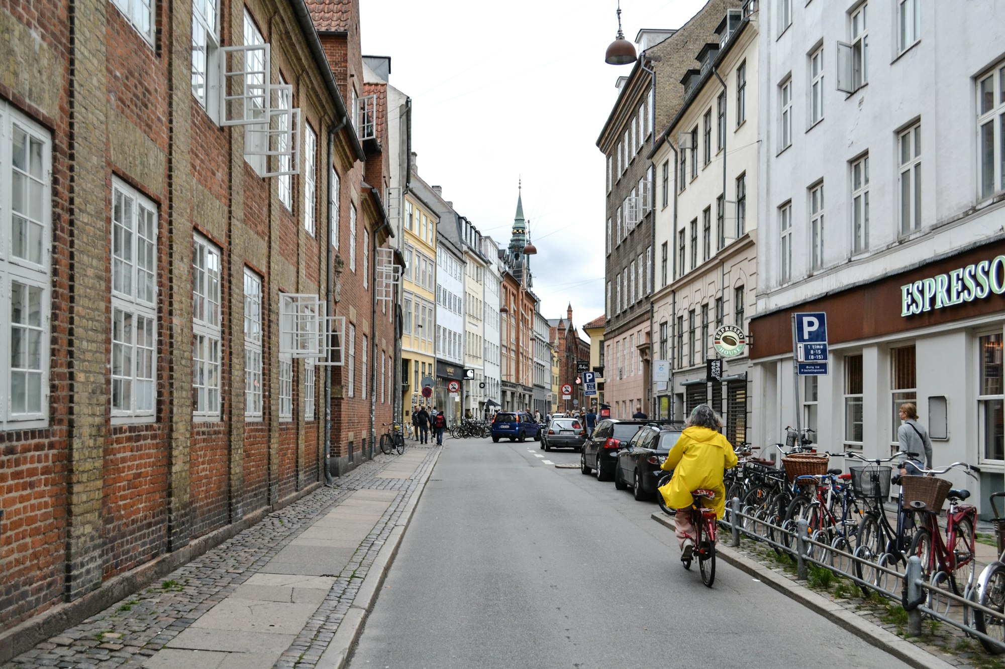 A cyclist in a yellow raincoat riding down a cobbled Copenhagen street lined with bicycles and cafés