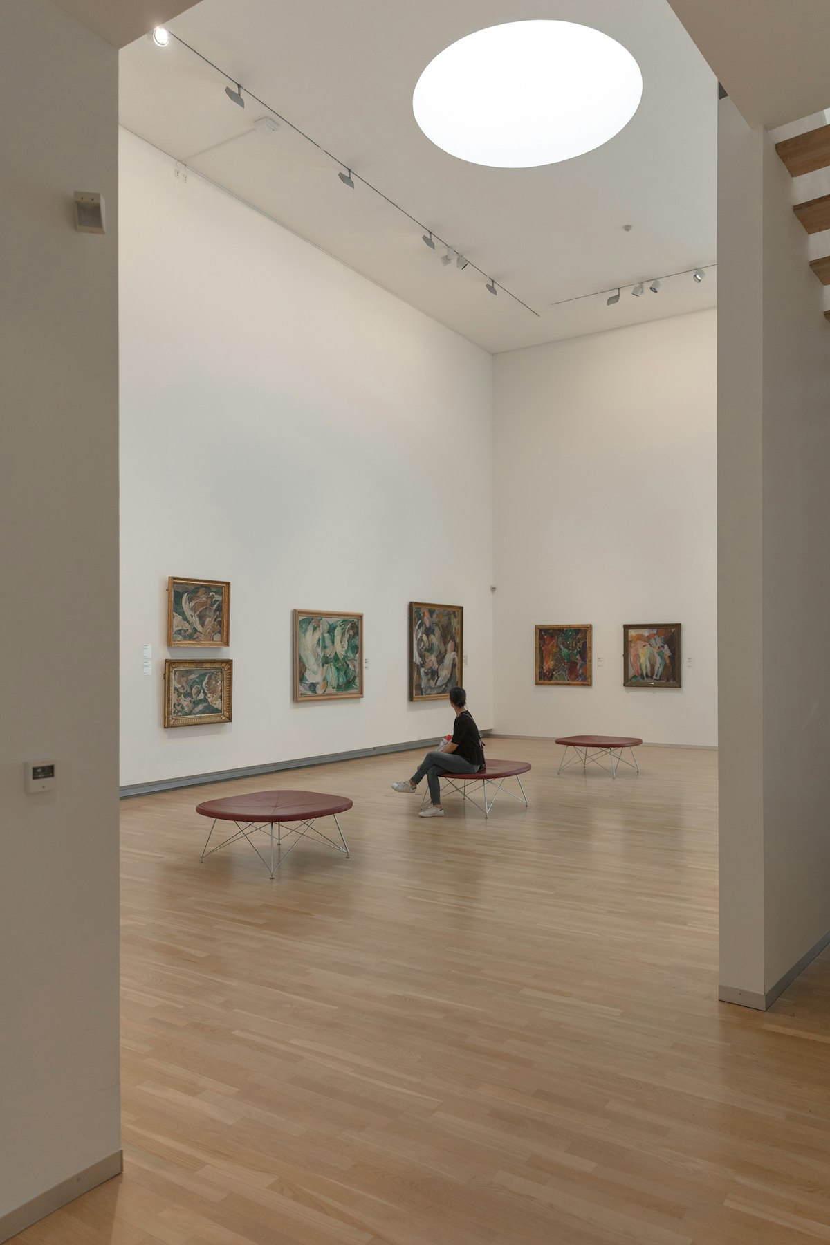 A light-filled gallery with pale wood floors and a visitor seated before a wall of paintings